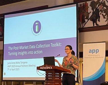 Image of a woman standing at a podum in front of an I2I presentation on the PMDC. A banner that says, 'app' is at the right of the image.